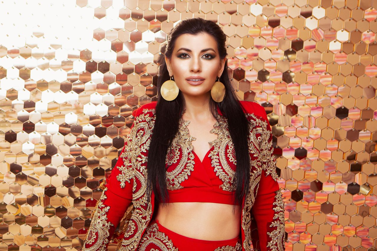 Attractive female wearing red costume and round earrings looking at camera while standing in light studio on background with golden sequins